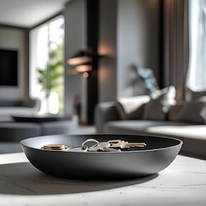 May include: A black, round decorative bowl on a white surface, containing keys and coins. The bowl has a textured surface and is in focus, with a blurred living room background. The keys are silver and gold.