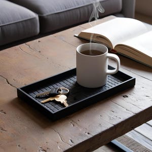 May include: A black rectangular tray with a textured surface holds a white mug with a hot beverage, keys, and a car key. Steam rises from the mug. An open book rests on a wooden table, near a gray sofa.