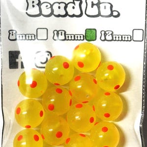 Op de afbeelding: Een zak met 12 mm gele kralen met rode stippen. De kralen zijn doorschijnend en hebben een glanzende afwerking. De zak is gelabeld met "Ruddy Roe Bead Co."