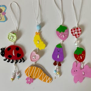 May include: A collection of handmade wooden charms featuring various designs. The charms include a ladybug, a slice of salmon, a yellow banana, a pink heart, a purple eggplant, a strawberry, a checkered heart, and a pink bunny. Each charm is painted with bright colors.