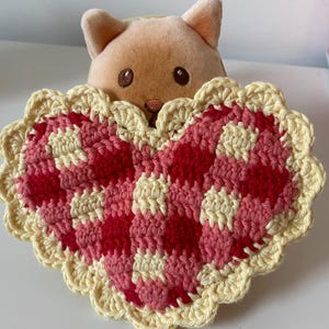 May include: A crocheted heart-shaped coaster with a scalloped edge. The coaster is made with yellow and pink yarn and is sitting in front of a plush toy.