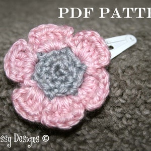 May include: A pink and gray crocheted flower hair clip. The flower has five petals and a center made of gray yarn. The clip is attached to the back of the flower.