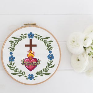 May include: A circular wooden embroidery hoop containing a cross-stitch pattern of a Sacred Heart with a brown cross, yellow and orange flames, and a green crown of thorns, surrounded by a wreath of green leaves and small blue flowers.