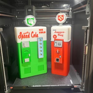 May include: Two 3D-printed vending machine replicas. The green one features the text 'Drink Speed Cola' and 'ICE COLD' with a white hand icon topper. The red one features the text 'Drink Jugger-Nog' and '10c' with a white lightning bolt icon topper.