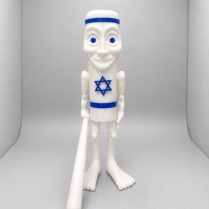 May include: A white plastic figurine with a cylindrical body, a smiling face, and blue eyes. The figure features a blue Star of David on its chest, blue horizontal bands at the top and bottom, and holds a long white cylindrical rod in its right hand.