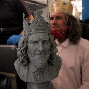 May include: A gray 3D-printed bust of a person with long wavy hair, wearing a Burger King crown and a bandana around the neck. The sculpture features a collared shirt and is mounted on a square base, capturing the likeness of the person behind it.