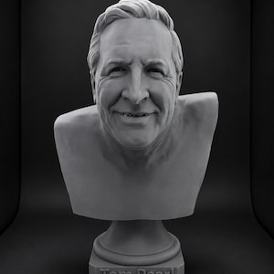 Pode incluir: Um busto escultural cinza monocromático representando uma pessoa com cabelo curto, um sorriso suave e traços faciais visíveis. A escultura está montada sobre uma base quadrada com o nome Tom Pearl em relevo na parte frontal.