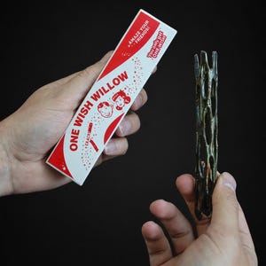 May include: A rectangular white box with red accents labeled ONE WISH WILLOW, featuring cartoon faces and the text AMAZE YOUR FRIENDS! and You only get ONE WISH. Beside it is a dark brown, cylindrical, porous, and hollow wooden stick.