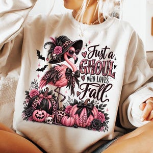 May include: A cream-coloured crewneck sweatshirt featuring a graphic of a pink flamingo wearing a black hat and sunglasses, surrounded by pink and black pumpkins, roses, and bats, with the text Just a GHOUL WHO LOVES Fall printed in black and pink.