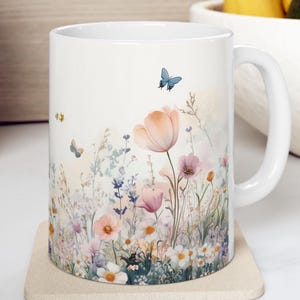 Könnte beinhalten: Weiße Keramiktasse mit einem Blumendesign mit rosa und weißen Blumen, grünen Blättern und einem blauen Schmetterling. Das Design befindet sich auf der Vorderseite der Tasse und verläuft um die Seite.