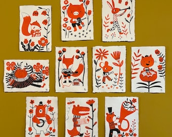 May include: A collection of ten whimsical art prints featuring orange and black animal illustrations. Each print showcases a different animal, such as a fox, rabbit, and bear, with floral accents. The prints are on textured, off-white paper, creating a folk art aesthetic.