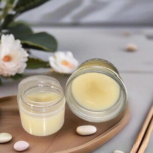 Puede incluir: Dos tarros de vidrio transparente con tapas que contienen un bálsamo o crema de color amarillo pálido. Los tarros están colocados en una bandeja de madera con algunas piedras blancas pequeñas. Dos palitos de madera están en el fondo.