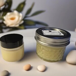 Pode incluir: Dois frascos de vidro com tampas contendo um creme verde claro e um creme amarelo. Os frascos estão sobre uma superfície branca com pequenas pedras decorativas.