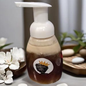 Peut inclure: Une bouteille en verre brun avec un distributeur à pompe blanc remplie d'un liquide couleur miel. La bouteille porte une étiquette ronde avec une silhouette noire d'une tête de femme portant une couronne et le texte "Queen's Raw Honey Foaming Face Wash Net Wt 5.8 oz."