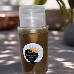 Puede incluir: Una botella de vidrio transparente con una etiqueta negra y dorada que dice "Queen's Rose Water Toner" con un gráfico de corona. La botella contiene un líquido marrón y tiene una tapa de plástico transparente.