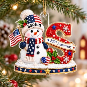 Puede incluir: Un adorno navideño con un muñeco de nieve que lleva un gorro y una bufanda con patrones de la bandera estadounidense y sostiene una bandera. Al lado hay una letra S roja grande con el texto 250th Anniversary, acebo y un corazón.