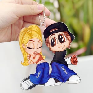 May include: A cartoon couple keychain with a girl with blonde hair wearing blue jeans with the name "Sophia" and a boy with black hair wearing blue jeans with the name "William".