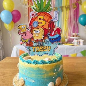 May include: A round birthday cake with blue and yellow ombre frosting, decorated with white seashells and sand-like crumbs at the base. A large pineapple-shaped cake topper features SpongeBob SquarePants characters, the number 5, and the name TOBBY.