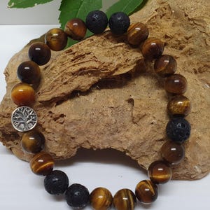 May include: A bracelet made of tiger's eye and black lava stone beads. The bracelet has a silver-coloured charm with a tree design. The tiger's eye beads are brown and gold, and the lava beads are dark grey.