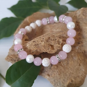 May include: A bracelet with pink, white, and silver beads. The bracelet has pink and white round beads, white textured beads, and silver spacers with clear rhinestones. The bracelet is displayed on a brown surface with green leaves. The bracelet's diameter is approximately 6 cm.