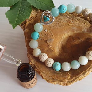 May include: A bracelet made of turquoise, white, and beige beads, with a silver heart charm. A small brown bottle with a dropper is next to the bracelet. The bottle has a label that reads "pepette essential oil".