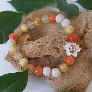 May include: A bracelet with a mix of orange, yellow, and white beads. The bracelet has a silver-coloured flower charm with a double ring design. The beads are strung on a stretchy cord, suitable for various wrist sizes.