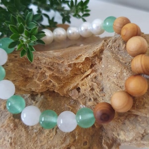 May include: A bracelet with a combination of white, green, and wooden beads. The bracelet is presented on a textured, brown surface, with green foliage in the background. The beads are round and of varying sizes.