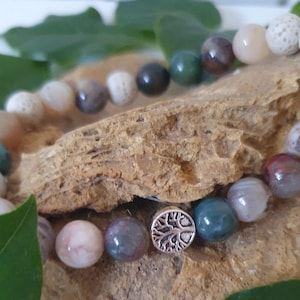 May include: Close-up of two beaded bracelets with a variety of stone beads in shades of white, grey, green, and pink. One bracelet features a silver charm with a tree design. The bracelets are displayed on a textured, brown surface.