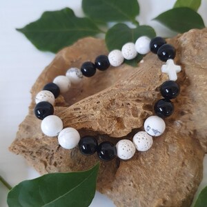 May include: A bracelet made of black and white round beads, with a white cross-shaped bead. The bracelet is presented on a textured, brown surface, with green leaves. The beads are approximately 1 cm in diameter. A religious-themed accessory.