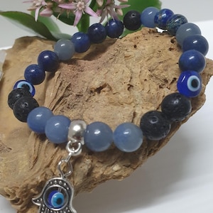 May include: A bracelet with blue and grey gemstone beads, black lava beads, and a silver Hamsa hand charm with an evil eye. The bracelet is displayed on wood, with pink flowers in the background.