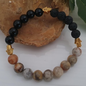May include: A bracelet made with black onyx, lava rock, and patterned stone beads. Gold-coloured Hamsa hand charms are featured. The bracelet is strung on an elastic cord.