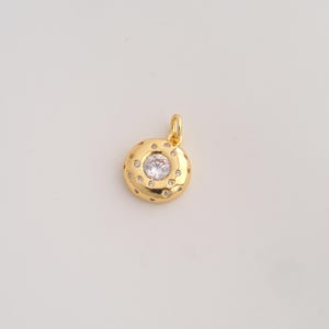 May include: A circular gold pendant featuring a central clear round-cut gemstone surrounded by multiple smaller embedded clear stones. The pendant has a polished gold finish and includes a small gold jump ring at the top for attachment.