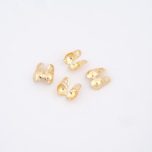 May include: Several small, gold-colored jewelry findings are scattered on a white surface. Each finding has a unique shape, with a rounded base and a loop for attaching to other components. The findings appear to be made of metal and have a shiny finish.