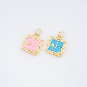 Könnte beinhalten: Zwei goldfarbene Anhänger mit Fensterdesign. Einer hat einen rosa Hintergrund, der andere ist blau. Beide haben einen quadratischen Rahmen mit dekorativem Rand und eine kleine Schlaufe für eine Kette. Die Fensterscheiben sind sichtbar.
