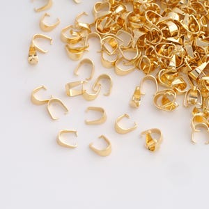 May include: A selection of small, gold-coloured jewellery findings, including bail connectors and clasps, scattered on a white surface. The findings are in various shapes, with a shiny, metallic finish, ideal for jewellery making.
