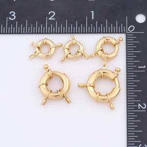 May include: Five gold-toned metal jump rings of varying sizes. The rings are round and have a decorative design. The rings are arranged in a row on a white surface.