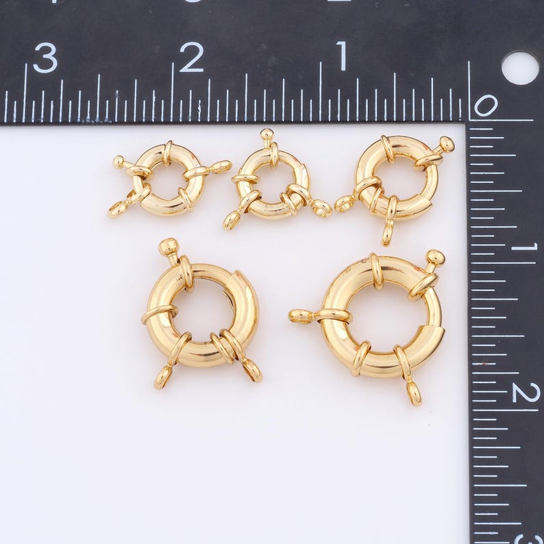 May include: Five gold-toned metal jump rings of varying sizes. The rings are round and have a decorative design. The rings are arranged in a row on a white surface.