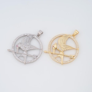 May include: Two silver and gold pendants featuring a mockingjay bird with a red eye and an arrow through it, surrounded by a circle of clear rhinestones.