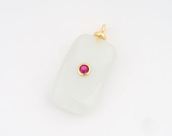 Puede incluir: Un colgante rectangular con una fianza dorada y una pequeña gema roja redonda engastada en un bisel dorado. El colgante es de un color blanco suave y opaco.
