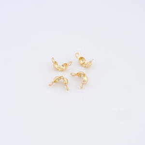 May include: Four small, gold-colored crimp bead findings, used in jewelry making. Each bead is open, showing the interior, and has a small loop for attaching to other components. The beads are scattered on a white surface.