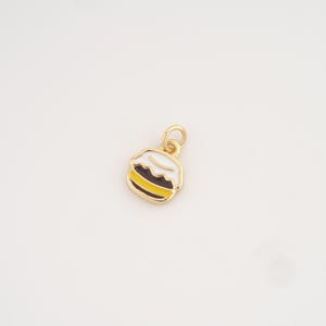 Puede incluir: Un dije dorado con forma de postre, posiblemente un macaron. El dije presenta una parte superior blanca, un centro marrón oscuro y una parte inferior amarilla, todo delineado en negro. El dije tiene un pequeño lazo para sujetarlo a un collar o pulsera.
