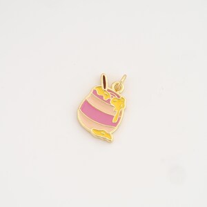Puede incluir: Un dije dorado en forma de tarro de miel con esmalte rosa y amarillo. El dije tiene un pequeño lazo en la parte superior para sujetarlo a un collar o pulsera. El tarro de miel tiene rayas rosas y beige.