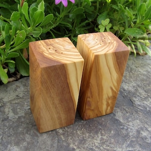 Op de afbeelding: Een set van twee houten zout- en peperstrooiers gemaakt van olijfhout. De strooiers zijn rechthoekig en hebben een natuurlijke houtkleur. Ze hebben kleine gaatjes aan de bovenkant om zout en peper te strooien.
