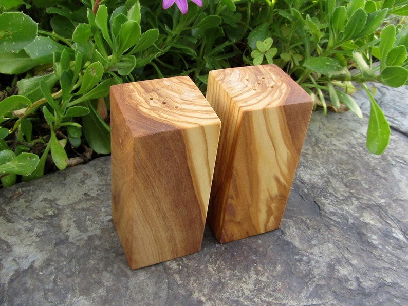 Op de afbeelding: Een set van twee houten zout- en peperstrooiers gemaakt van olijfhout. De strooiers zijn rechthoekig en hebben een natuurlijke houtkleur. Ze hebben kleine gaatjes aan de bovenkant om zout en peper te strooien.