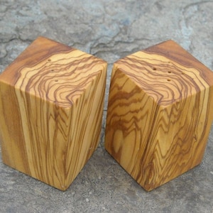 May include: A set of two wooden salt and pepper shakers made from olive wood. The shakers are square with rounded edges and have a natural wood grain pattern.