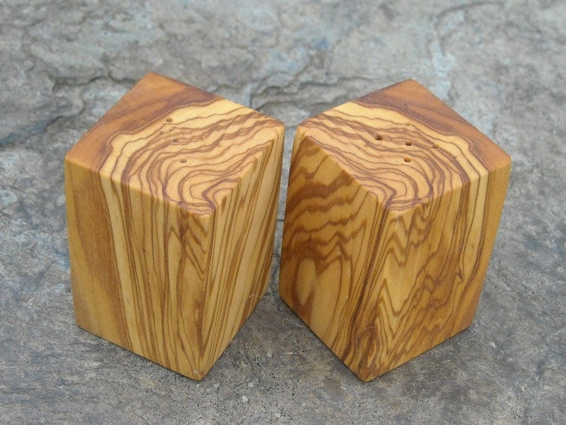 May include: A set of two wooden salt and pepper shakers made from olive wood. The shakers are square with rounded edges and have a natural wood grain pattern.