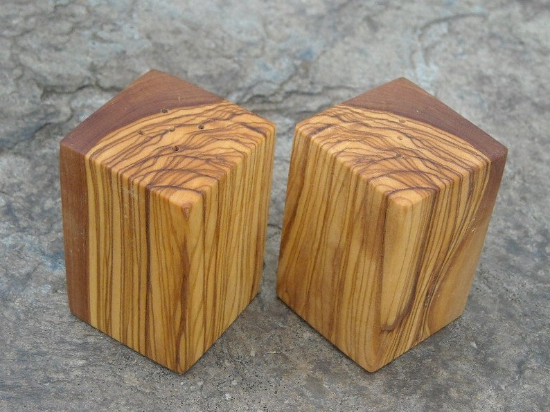 May include: Two wooden salt and pepper shakers made from olive wood. The shakers are square shaped and have a natural wood finish.