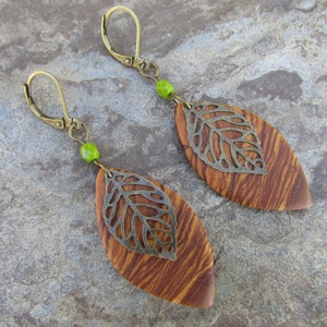 May include: A pair of earrings with a wood and metal leaf design. The leaves are brown and have a silver metal overlay. The earrings are attached to a gold hook with a green bead.