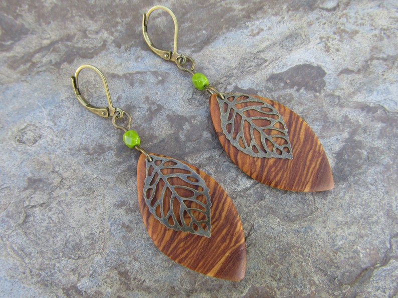 May include: A pair of earrings with a wood and metal leaf design. The leaves are brown and have a silver metal overlay. The earrings are attached to a gold hook with a green bead.