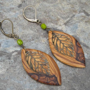 May include: A pair of olive wood earrings with a leaf design. The earrings are made of wood with a dark brown and light brown pattern. The leaves are made of metal and are a gold color. The earrings have green beads on the hooks.